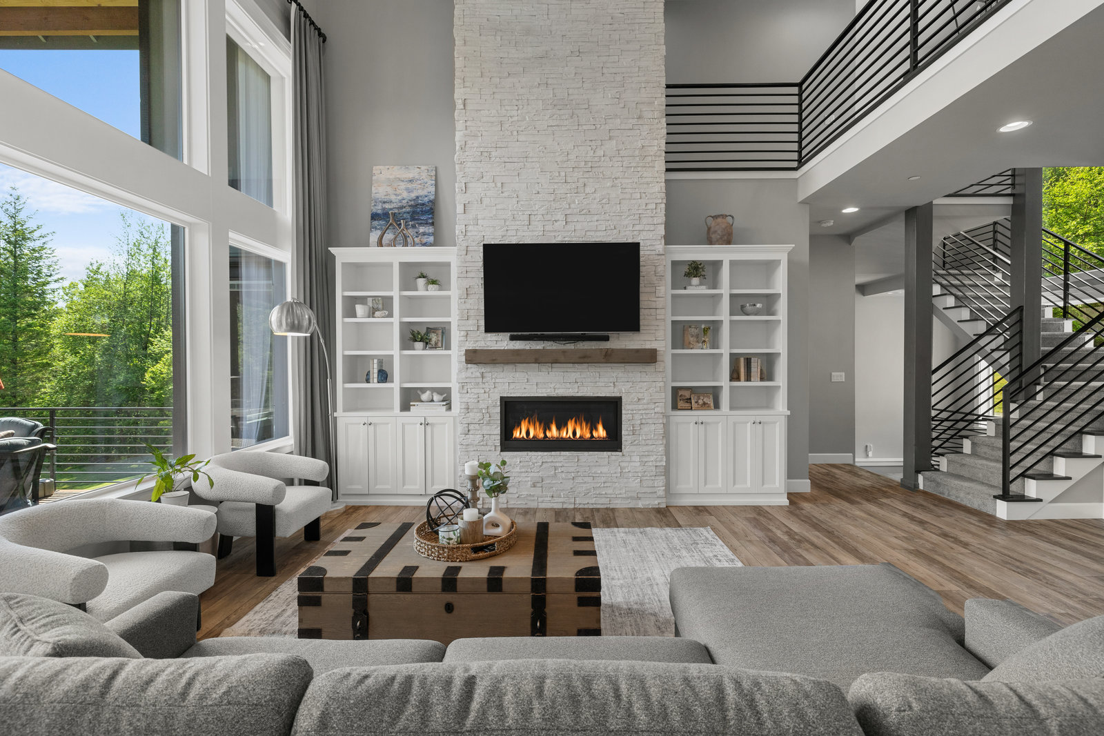 The dramatic two-story living room showcases a floor-to-ceiling stone fireplace, built-in bookshelves, Restoration Hardware designer lighting, and elegant custom drapery. A semi-floating staircase with sleek custom handrails leads to the upper-level bedrooms and office